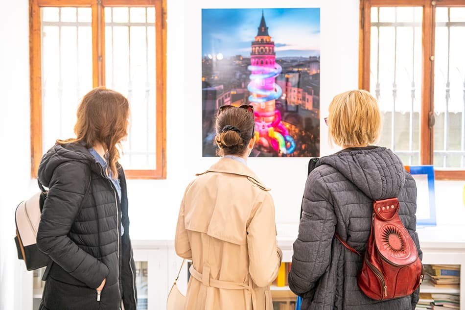 Three visitors seen from behind in a white gallery room with wooden-framed windows, looking at a large print of Istanbul's Galata Tower wrapped in a rainbow spiral of light at dusk.